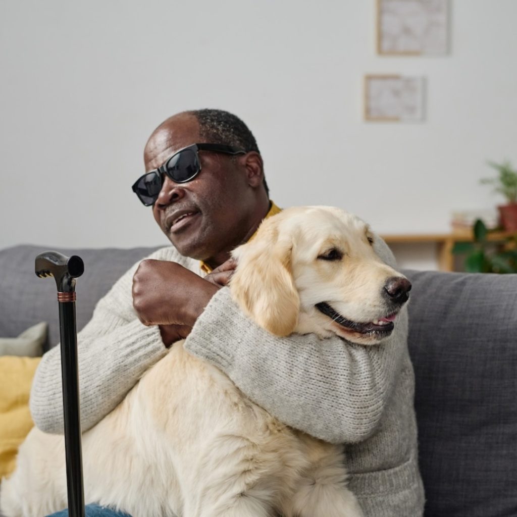 Senior man in glasses with bad sight embracing his individual guide dog while sitting on sofa at home; fair housing month
