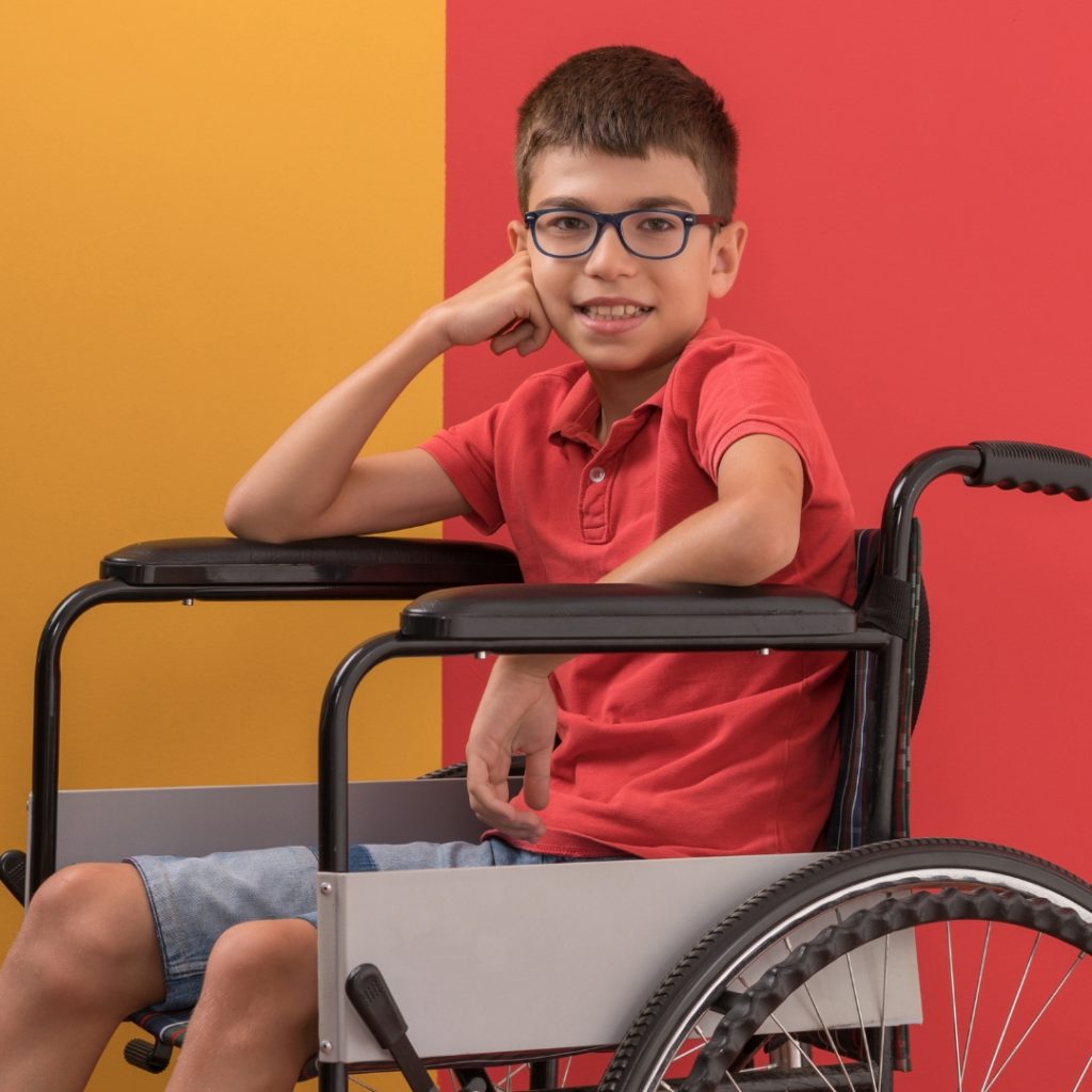 A boy wearing glasses and a red polo shirt sits in a wheelchair, leaning his head on his hand, against a background split between orange and red; fair housing month